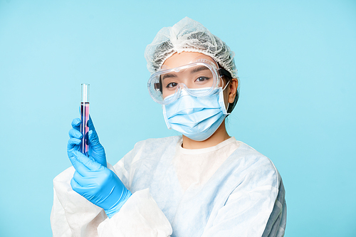 Asian nurse or lab worker in personal protective equipment, showing test sample tube, standing in medical face mask over blue background.