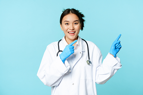 Happy smiling nurse, asian female doctor pointing right, showing promo, clinic advertisement, standing in medical uniform over blue background.