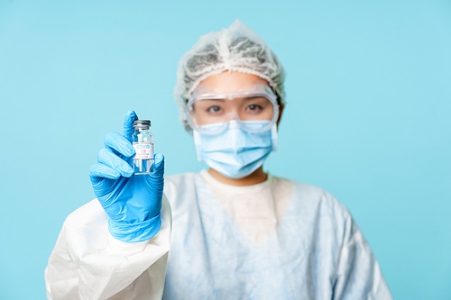 Covid-19 vaccination concept. Asian female nurse or doctor in personal protective equipment, showing coronavirus vaccine bottle, standing over blue background.