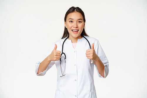 Happy smiling asian woman doctor, showing thumbs up in approval, satisfied with patient condition, approve and like, standing over white background.
