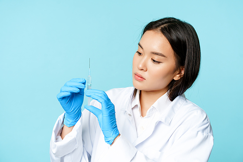 Image of asian female nurse, doctor in uniform and gloves, holding syringe, standing over blue background.