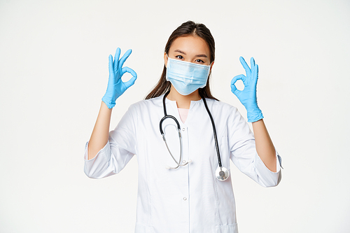 Smiling nurse, female asian doctor in rubber gloves, medical face mask shows okay, OK signs, stands against white background.