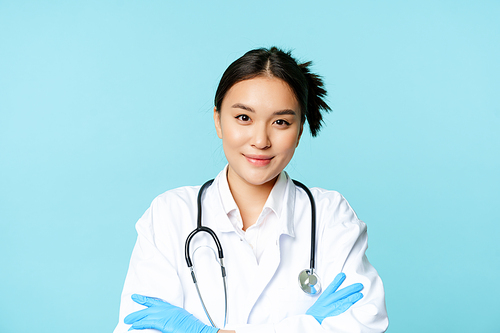 Smiling asian woman doctor, nurse in gloves and clinic uniform, cross arms and looking determined, blue background.