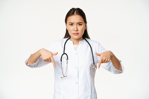 Disappointed asian female doctor pointing fingers down, frowning and looking concerned, showing bad product, standing over white background.