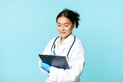 Asian female nurse or doctor writing down on clipboard with pen, smiling while examining patients, standing in uniform over blue background.