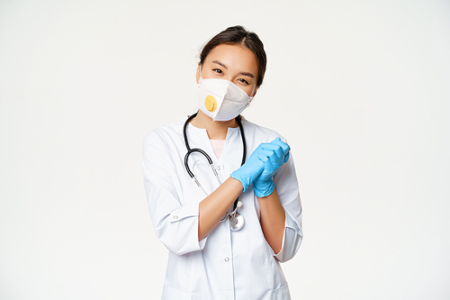 Smiling cute asian female doctor, nurse in medical respirator and rubber sterile gloves, looking pleased at camera, standing in healthcare worker uniform, white background.