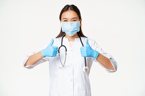 Healthcare and covid-19 preventive measures concept. Asian physician in face mask and rubber gloves, shows thumbs up, stands in uniform over white background.