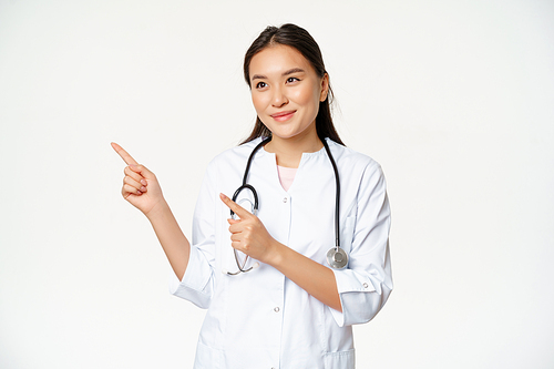 Pleasant smiling female doctor in uniform, pointing fingers and looking left with caring, friendly smile, standing over white background.