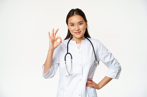 Very good. Smiling confident asian female doctor, showing okay, ok sign in approval, confirm smth, saying yes, give approval, standing in medical uniform, white background.