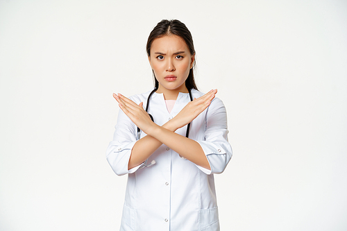 Serious asian woman doctor prohibit patient behaviour, showing stop, cross arms gesture, frownign displeased, disapprove smth bad, standing over white background.