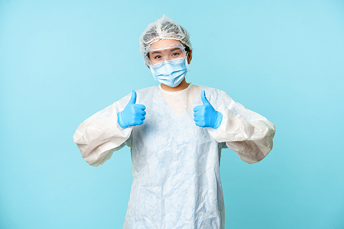Optimistic asian nurse or doctor, wearing personal protective equipment, showing thumbs up, campaign from coronavirus prevention, blue background.