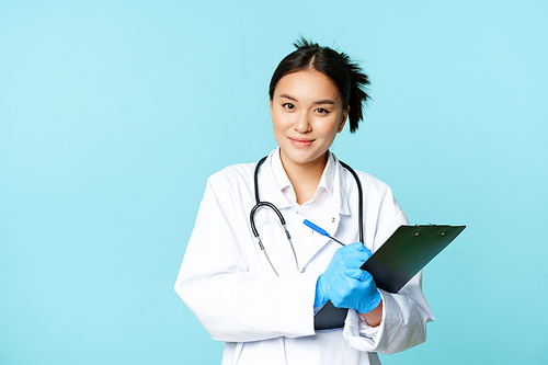 Smiling asian medical worker, female physician writing down patient info, holding pen and clipboard, standing in uniform over blue background.