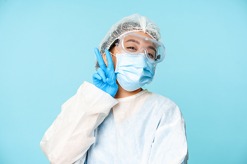 Happy, kawaii asian female doctor, nurse in personal protective equipment, showing v-sign peace gesture near face mask, blue background.