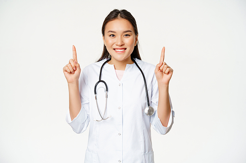 Smiling asian woman doctor, wearing medical robe, pointing fingers up at advertisement, showing promo, provide healthcare information, white background.