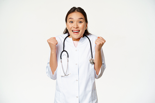 Enthusiastic female asian doctor celebrating, looking hopeful and happy at camera, triumphing, standing in medical uniform against white background.