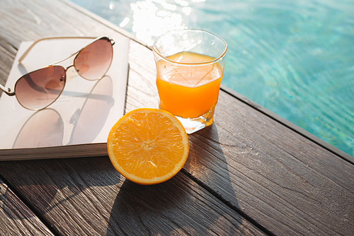 Orange fresh juice, book and sunglasses near pool