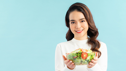 Portrait of a happy playful girl eating fresh salad from a bowl and winking isolated over blue background