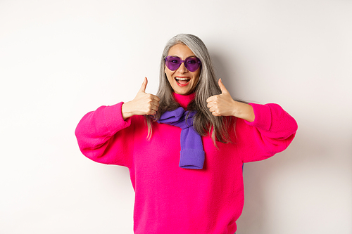 Shopping concept. Stylish asian senior woman in sunglasses and trendy outfit, showing thumb-up in approval, recommending shop, standing over white background.