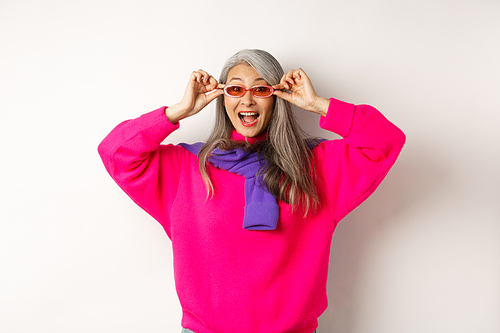 Fashionable asian senior woman trying new sunglasses, looking amazed and cheerful at camera, standing in trendy sweater against white background.