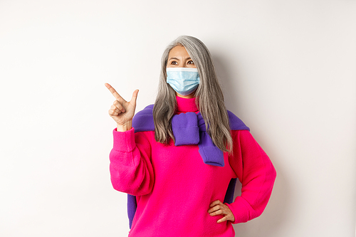 Covid-19, quarantine and lockdown concept. Stylish asian grandmother in face mask pointing and looking at upper left corner, showing logo and smiling, white background.