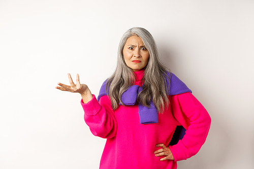 Angry and confused asian senior woman spread hand sideways and staring at camera puzzled, standing in pink sweater against white background.