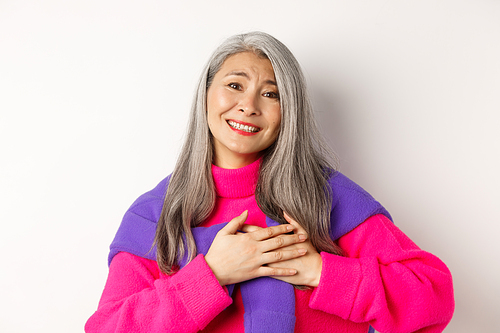 Close up of stylish asian mother holding hands on heart, looking touched and heartfelth, saying thank you, smiling grateful, standing over white background.