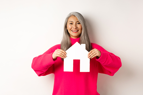 Real estate. Beautiful asian grandmother showing paper house and smiling happy, standing in pink sweater over white background.