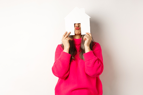 Real estate. Funny asian middle-aged woman hiding face behind paper house model and showing puckered lips, having fun, standing over white background.