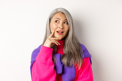 Close up of beautiful asian elderly woman in stylish clothes, making plans, looking aside and thinking, standing over white background.