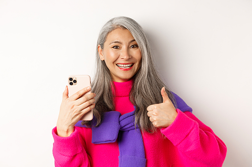 Online shopping. Close up of fashionable asian senior woman showing thumbs-up, using smartphone and approve something, standing over white background.