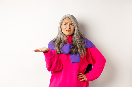 Funny asian mother with grey hair complaining, shrugging and looking left confused, pointing hand at something strange, standing over white background.