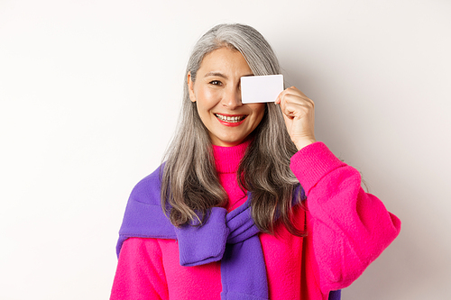 Shopping concept. Stylish asian senior woman smiling and showing plastic credit card, paying contactless, standing over white background.