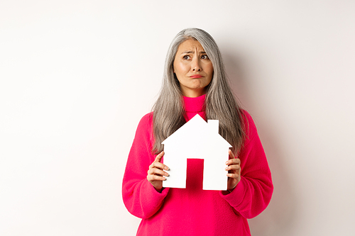 Real estate. Sad asian senior woman dreaming about own property, holding paper house model and grimacing upset, looking at upper left corner, white background.