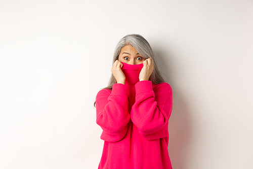 Portrait of funny asian grandmother hiding face in sweater collar, peeking at camera silly, standing over white background.