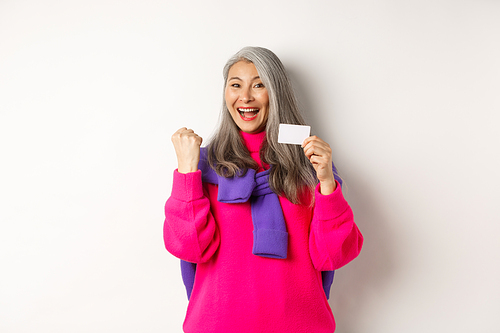 Shopping concept. Cheerful asian senior woman winning prize from bank, showing fist pump gesture and plastic credit card, standing happy over white background.