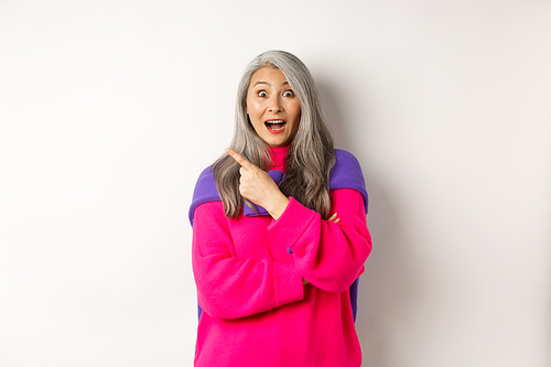 Portrait of impressed asian lady checking out promo offer, pointing at upper left corner and staring at camera amazed, standing in trendy pink sweater over white background.