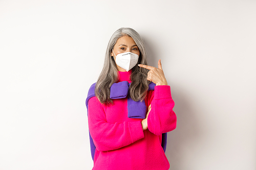 Covid, pandemic and social distancing concept. Fashionable asian senior woman wearing respirator, pointing at face mask and smiling, standing over white background.