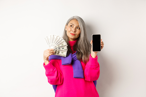 Fashionable asian senior woman showing money dollars and blank smartphone screen, demonstrate online shop, standing over white background.