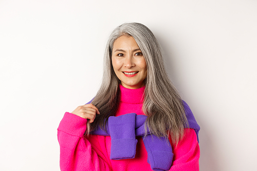 Close-up of beautiful asian senior woman in trendy pink sweater, smiling cheerful at camera, standing over white background.