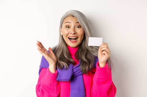 Shopping concept. Happy asian old aldy looking impressed and showing plastic credit card of her bank, standing over white background.