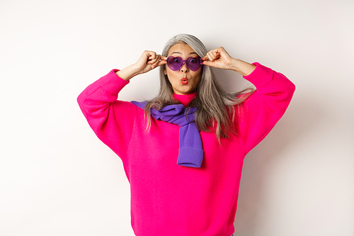 Fashionable asian senior woman in sunglasses and stylish pink sweater pucker lips, raising eyebrows and looking surprised at camera, white background.