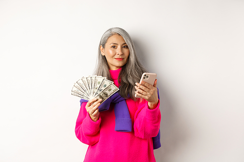 Modern asian senior lady holding money and smartphone, order something online, standing over white background.