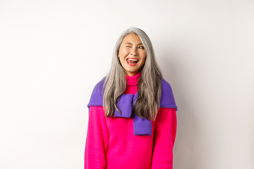 Funny and happy asian senior woman having fun, showing tongue and winking cheeky, looking left at logo, standing over white background.
