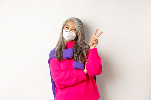 Covid, pandemic and social distancing concept. Cheerful and stylish asian senior woman wearing respirator from coronavirus, showing victory sign, standing over white background.