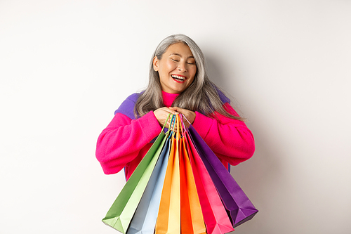 Fashionable senior asian woman shopaholic, hugging shopping bags and smiling joyful, buying with discounts, standing over white background.