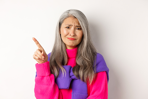 Judgy and skeptical asian senior woman grimacing displeased, pointing finger upper left corner unamused, standing over white background.