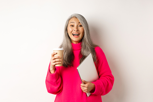 Happy asian senior female entrepreneur laughing, drinking coffee and holding laptop, standing in pink sweater over white background.