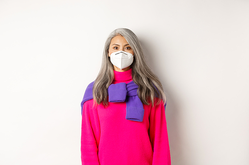 Covid, pandemic and social distancing concept. Stylish asian senior woman wearing respirator and looking at camera serious, standing over white background.