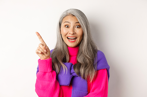 Close up of beautiful asian female model pointing finger at upper left corner, smiling and looking happy at camera, showing promo deal, white background.