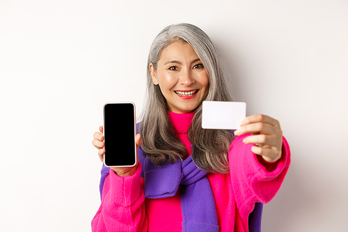 Online shopping. Close-up of stylish korean senior woman showing empty mobile screen and plastic credit card, smiling happy, standing over white background.